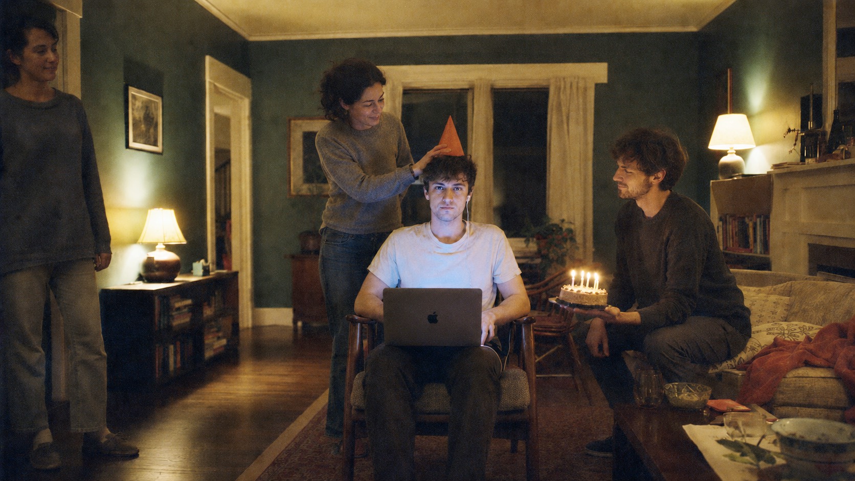 A man at his own birthday party, a laptop open on his lap, while a friend lowers a party hat onto his head and another waits beside him with the cake.